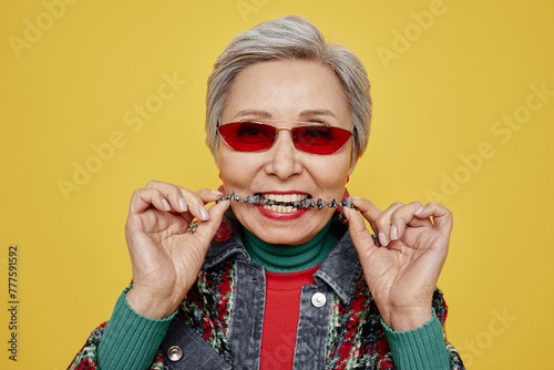 Aged woman in sunglasses and dopamine dressing biting her necklace with healthy teeth while posing in isolation on yellow background photo