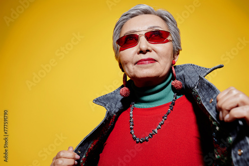 Cool senior woman with bright red lipstick and pullover looking through stylish sunglasses while posing on yellow background in studio