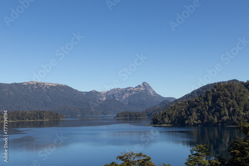 Majestic nature view of a huge clear blue lake reflecting the forest that surrounds it and the mountain range in the distance, in Lago Espejo, Argentina
