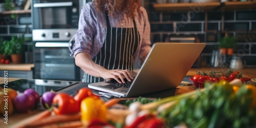 A woman in a kitchen filled with fresh vegetables using a laptop, likely searching for recipes or conducting a cooking class, suitable for culinary blogs or holiday meal prep.
