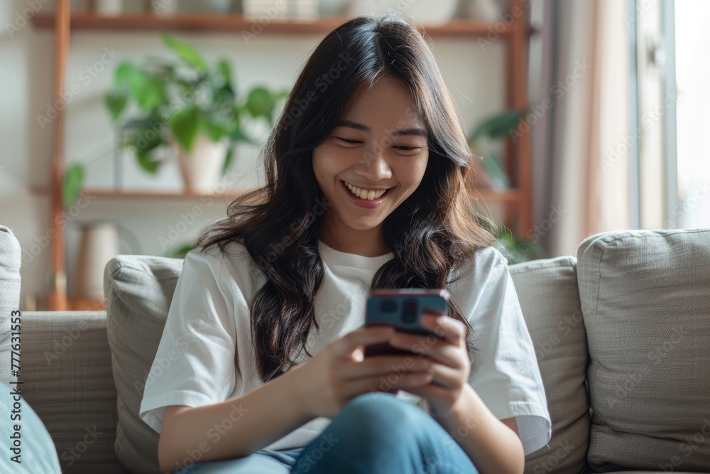 Happy young asian woman using mobile phone while sitting a couch at home.Happy relaxed young woman sitting on couch using cell phone, smiling lady laughing holding smartphone Happy young asian woman using mobile phone while sitting a couch at home.Happy relaxed young woman sitting on couch using cell phone, smiling lady laughing holding smartphone