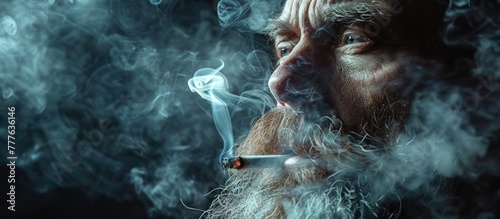 A bearded man is smoking a cigarette in the dark, illuminated by the glowing tip of the cigarette.