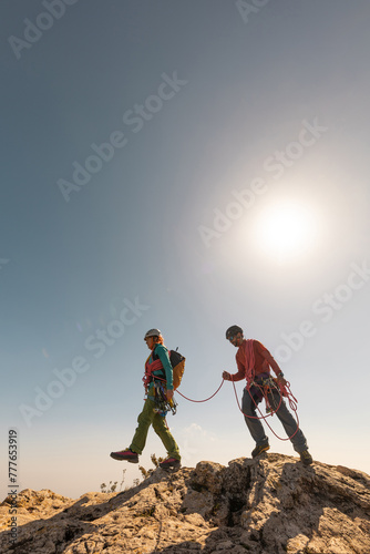 Two people are climbing a mountain together. One of them is wearing a red shirt. The sun is shining brightly in the sky