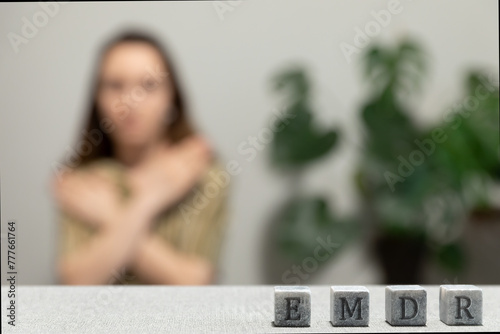 Letters EMDR written on grey stone cubes blocks. Female touching and tapping her shoulders in blurred background. Eye Movement Desensitization and Reprocessing psychotherapy treatment concept.