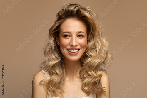 A young blonde woman with curly beautiful hair smiles on a beige background.