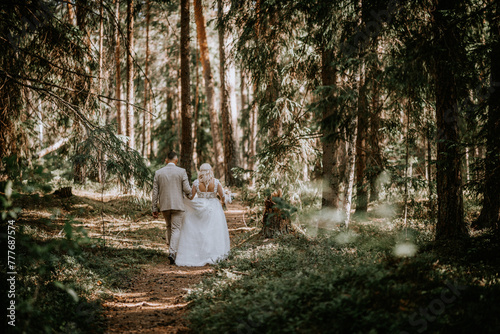 Valmiera, Latvia - Augist 13, 2023 - A bride and groom walk away hand in hand on a forest path, surrounded by tall pine trees and dappled sunlight. photo