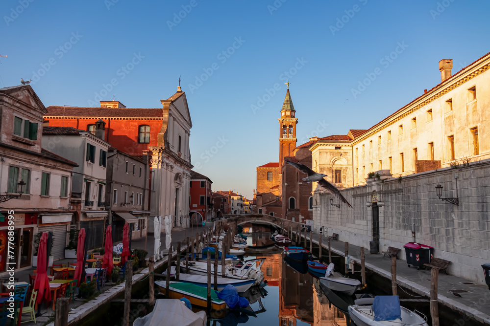 Fototapeta premium Church of Saint James Apostle with sunset view of canal Vena nestled in charming town of Chioggia, Venetian Lagoon, Veneto, Italy. Small boats floating in calm water creating romantic reflections