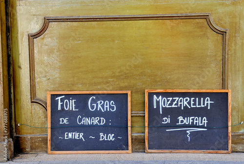 Two blackboards in France at a deli food shop. At the blackboard is written in Franch translated into English: duck foie gras the whole piece and buffalo mozzarella