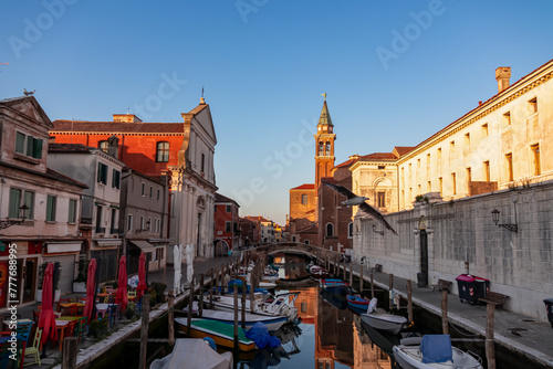 Church of Saint James Apostle with sunset view of canal Vena nestled in charming town of Chioggia, Venetian Lagoon, Veneto, Italy. Small boats floating in calm water creating romantic reflections