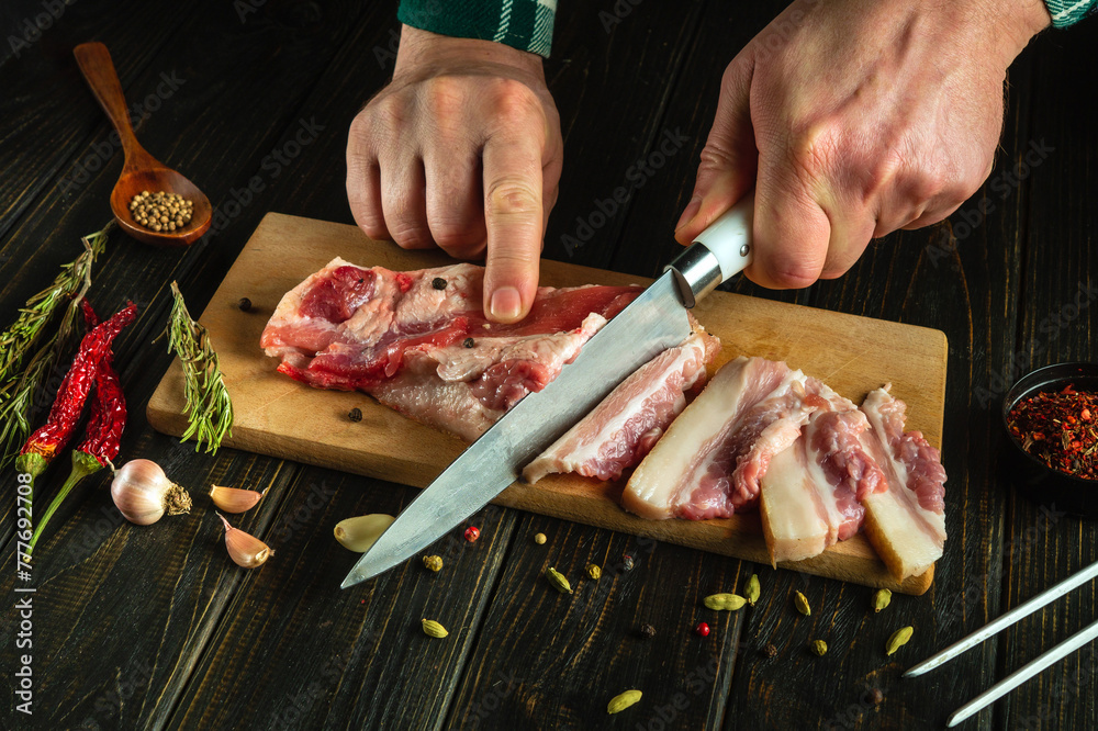 A butcher cuts lard with a knife on a kitchen board for preparing a grill or barbecue. Low key concept of cooking meat lunch.