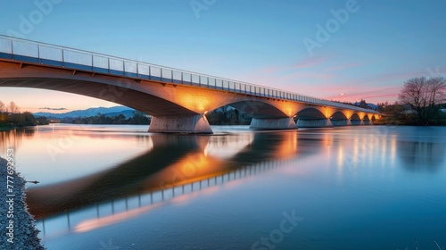 A bridge over a tranquil river at dusk, the structure illuminated by soft lighting against the backdrop of a sky transitioning from blue to pink, reflecting on the water below, highlighting the harmon © Muzammil Elahi