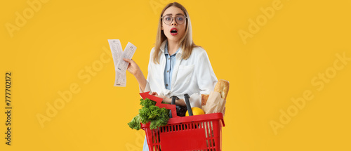 Shocked young woman with shopping basket of food and receipts on yellow background. Price rise concept photo