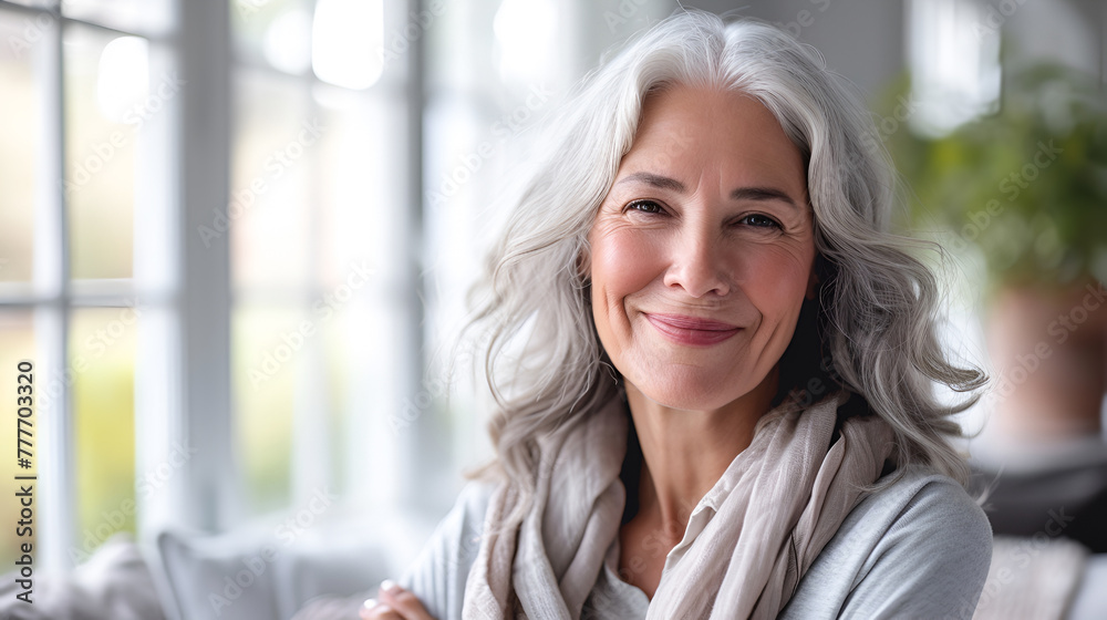 A smiling senior woman with long gray hair, a beige scarf, sits against the background of a window in a cozy, bright home interior with natural light. copy space