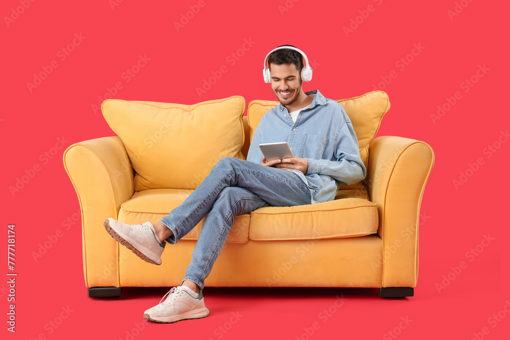 Handsome young happy man in headphones with tablet resting on sofa against red background Handsome young happy man in headphones with tablet resting on sofa against red background