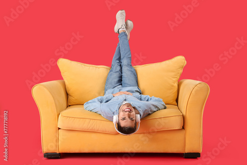Handsome young happy man in headphones resting on sofa against red background #777718192