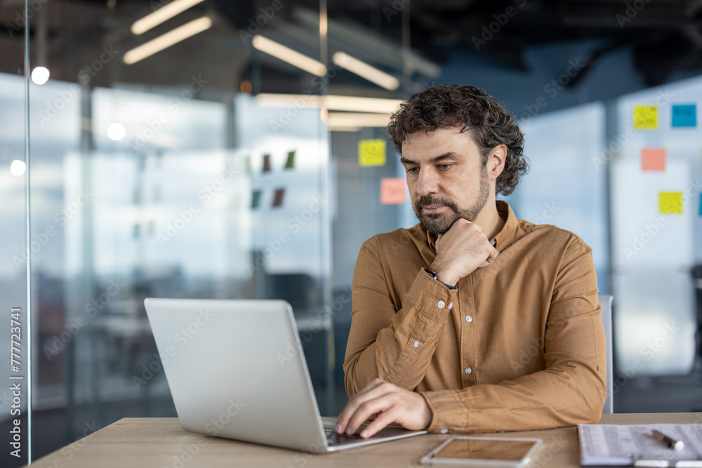 A serious Hispanic businessman deep in thought while working at his laptop in a modern office setting. A serious Hispanic businessman deep in thought while working at his laptop in a modern office setting.