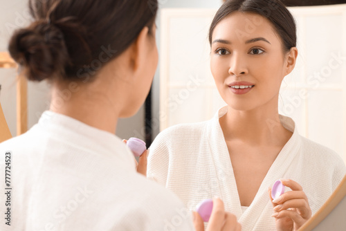 Young Asian woman with jar of facial cream near mirror in bathroom, closeup