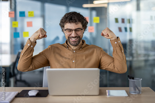 Joyous male professional in glasses celebrating a successful deal or achievement while working on his laptop in a modern office setting. © Liubomir