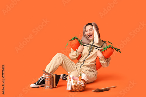 Happy female beekeeper in bunny ears with Easter basket, toy carrots and tools sitting on orange background photo