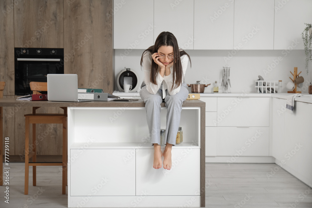 Tired young businesswoman sitting on table with laptop and office folders in kitchen Tired young businesswoman sitting on table with laptop and office folders in kitchen