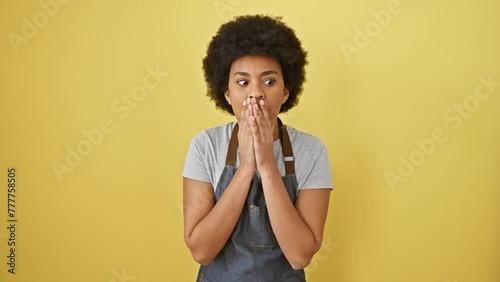 Astonished african american woman wearing apron, standing with a face of shock, scared and excited expression, mouth wide open in disbelief, isolated on yellow background photo