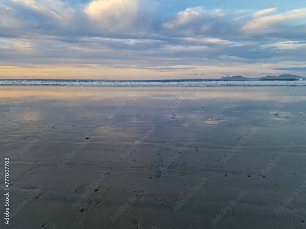Obraz premium Sunset on Famara Beach, Lanzarote, casts a mesmerizing palette of colors across the sky, blending hues of orange, pink, and purple against the backdrop of the Atlantic Ocean waves.