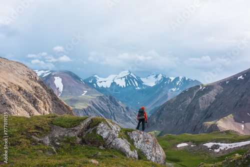 Man in vivid red jacket admire alpine scenery on green grassy hill among rocks near precipice edge. Guy with backpack on pass enjoying few big snowy pointy peaks. Three large snow peaked tops far away