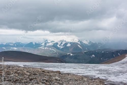 Dramatic panoramic view from big glacier to wide alpine valley and large snow-capped mountain range in rainy low clouds. Awesome vast landscape with high snowy mountains in rain under gray cloudy sky.