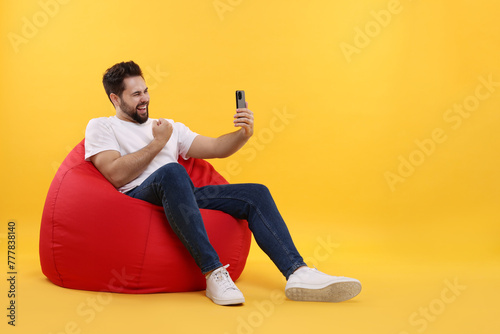 Happy young man using smartphone on bean bag chair against yellow background, space for text