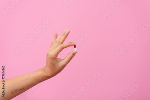 A female hand delicately holds a white pill between fingers against a pink isolated background, symbolizing healthcare, dietary supplements, and treatment for conditions depression and diseases.