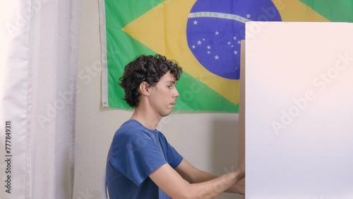 Jovem homem brasileiro escolhendo o seu candidato na urna eletrônica. Na parede, a bandeira do Brasil decora o local de votação. A imagem retrata as eleições brasileiras photo