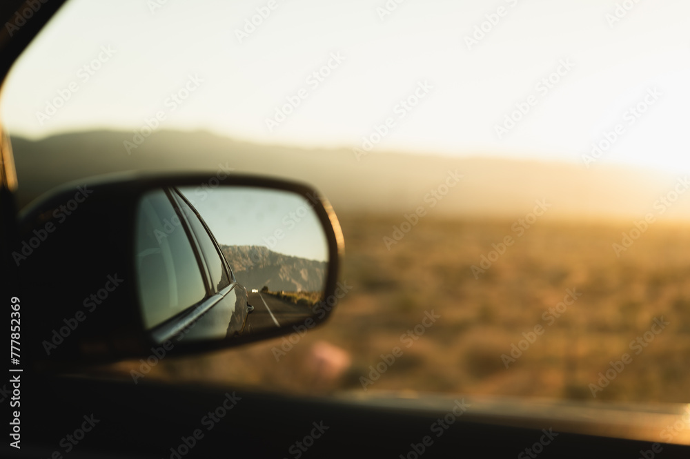 Reflection of car driving down a road as seen in side-view mirror against beautiful desert landscape at sunrise