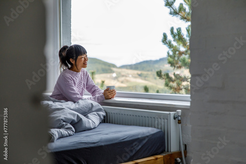 Woman enjoying coffee while relaxing by window on bed photo
