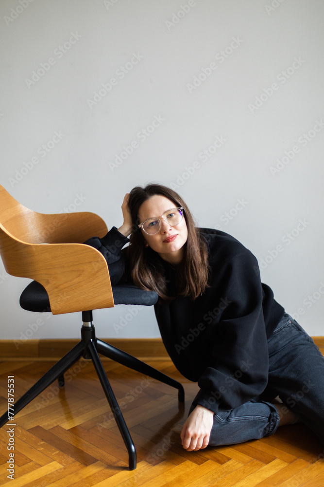 A woman sits on the floor leaning on a chair and  looks at the camera