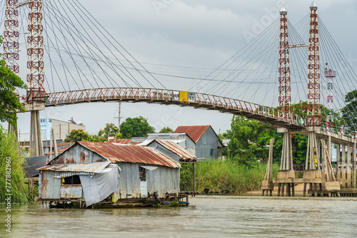 A rustic tin house floating on a river below a modern suspension bridge at Chau Doc in Vietnam