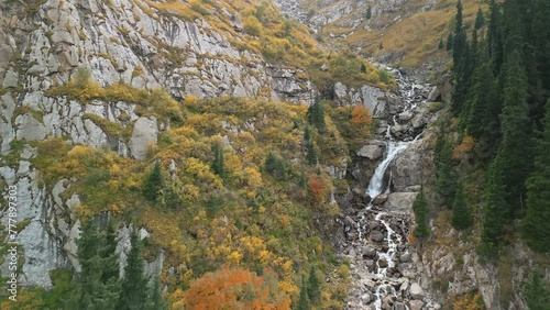 Barskoon Waterfall, Kyrgyzstan. Beautiful aerial drone view following the waterfall, autumn colours pine trees and mountains. Cinematic forward travelling shot. Famous adventure travel landmark. photo