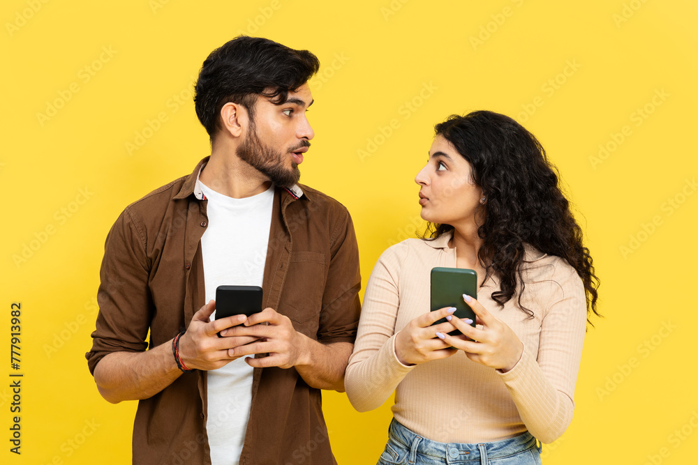 Curious Couple Looking At Each Other While Holding Smartphones Against A Yellow Background Curious Couple Looking At Each Other While Holding Smartphones Against A Yellow Background