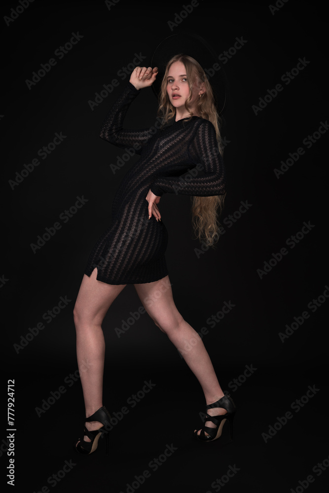 Full length portrait of woman is posing in front of black background. Blonde model with long hair is wearing black short tight knitted transparent dress, wide-brimmed felt hat and high heeled shoes Full length portrait of woman is posing in front of black background. Blonde model with long hair is wearing black short tight knitted transparent dress, wide-brimmed felt hat and high heeled shoes