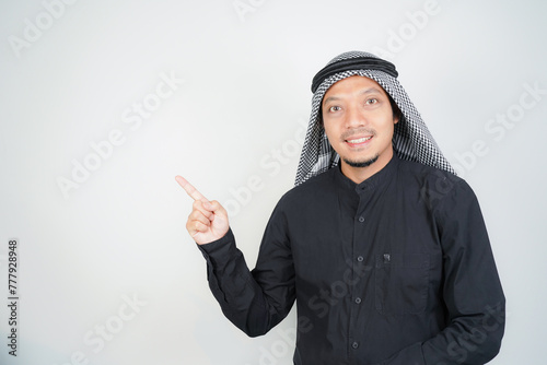 Cheerful Asian Muslim man wearing Arab turban sorban pointing hand finger at empty space on isolated photo