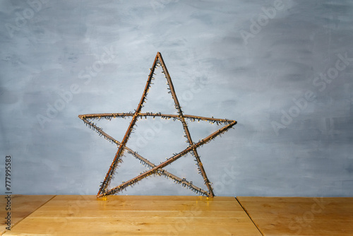 Decorative Twig Star with Lights Against Blue Wall. A handcrafted star made of twigs and adorned with small lights stands on a wooden floor against a textured blue wall.