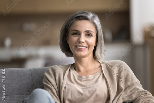 Close up portrait of smiling mature woman looking at camera, pose for photo, relax on cozy couch in living room, feels carefree on weekend alone indoors, pleasant happy lady enjoy leisure time at home © fizkes