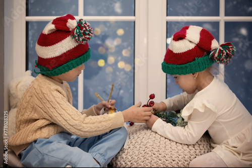 Little children brother and sister in Christmas hats play while sitting on a window with Christmas decor.