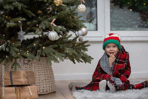 A happy little girl in a Santa Claus hat is wrapped in a warm plaid blanket by the Christmas tree.
