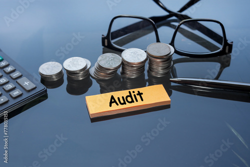Post-it note with the word AUDIT. Stacked coins, eyeglasses, and a calculator at the back. Business and financial audit concept