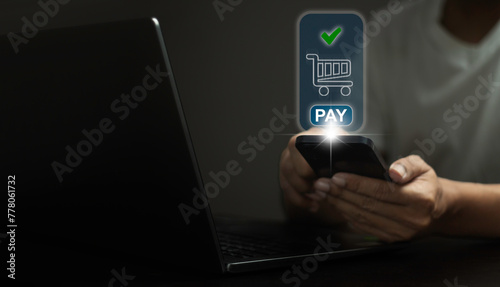 A person is using a laptop and a cell phone to pay for something. The laptop screen shows a shopping cart and the cell phone screen shows a check mark photo