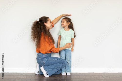 Young mom measuring her daughter's height holding hand above her head near white wall, full length, free space for text photo