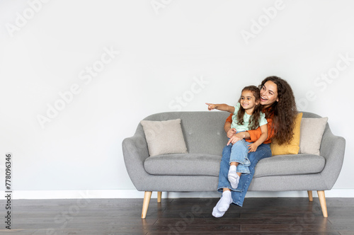 Mom and cute daughter pointing aside at free space while relaxing on sofa over white wall, showing space for advertisement