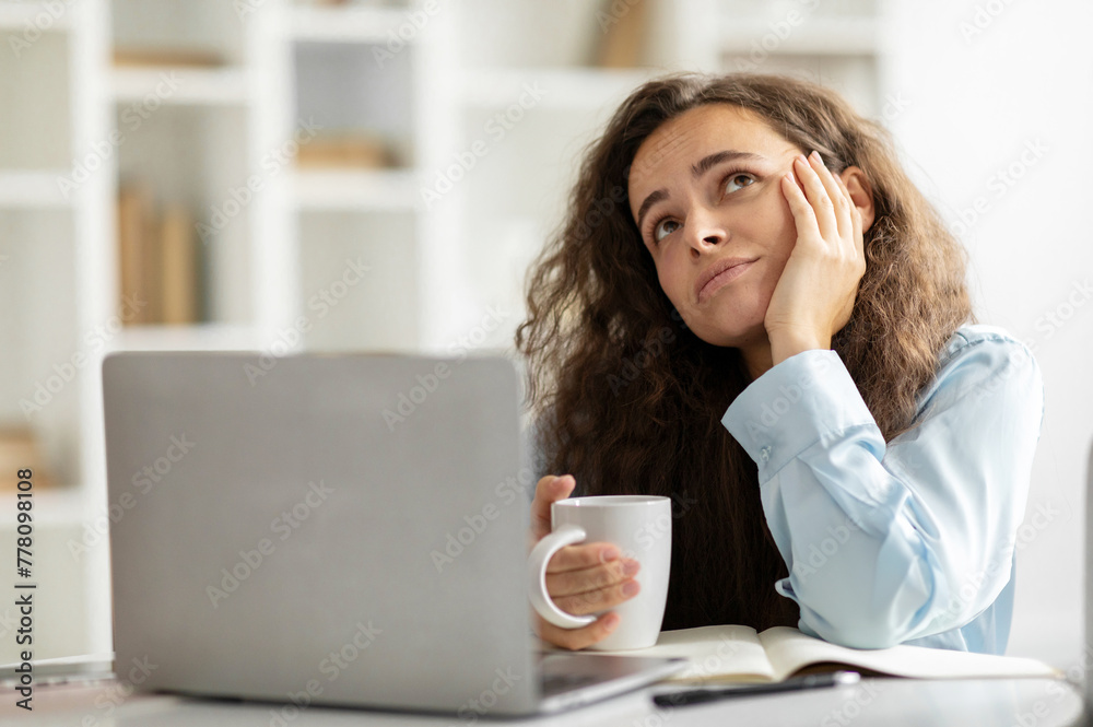 Pensive european woman sitting at desk with laptop and cup of coffee, thinking and looking up, dreaming about vacation Pensive european woman sitting at desk with laptop and cup of coffee, thinking and looking up, dreaming about vacation