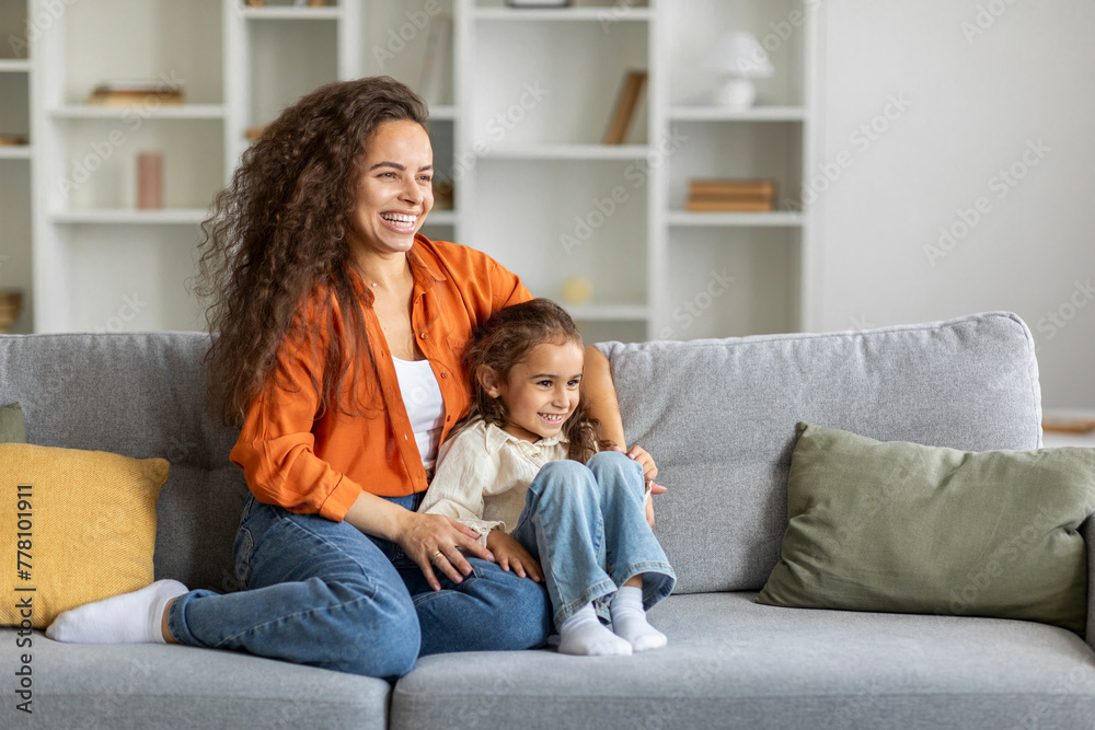Dreamy mother embracing small daughter, resting on couch and looking away, enjoy moments together at home, free space