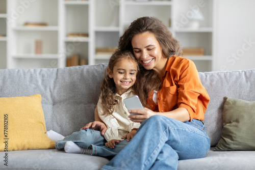 Cheerful mother and daughter using smartphone together, sitting on couch at home, shopping online on mobile phone and hugging